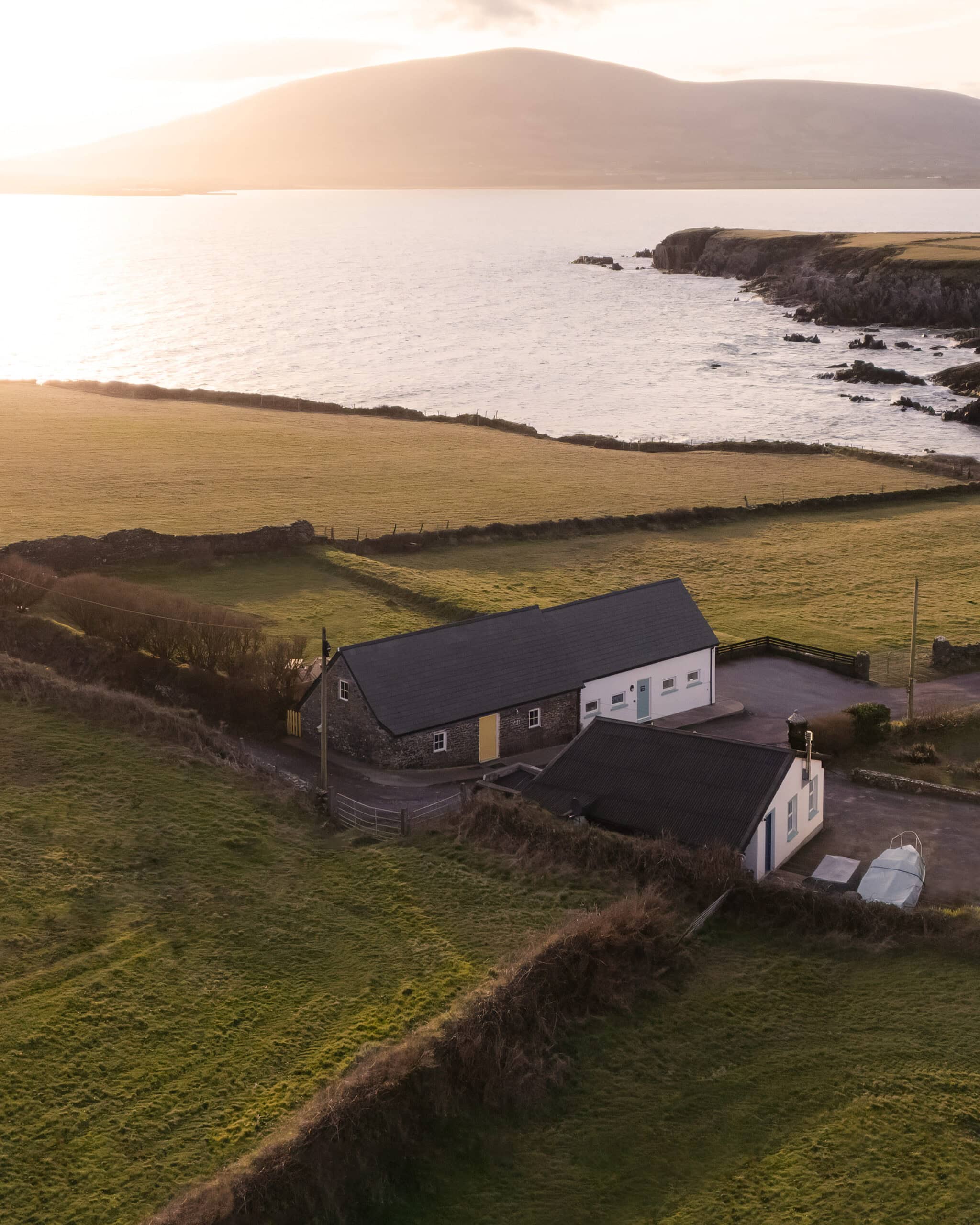 Aerial view of Pipers Cottage in Ballymore near Ventry Bay, Dingle, surrounded by green fields and overlooking the Atlantic Ocean at sunset.