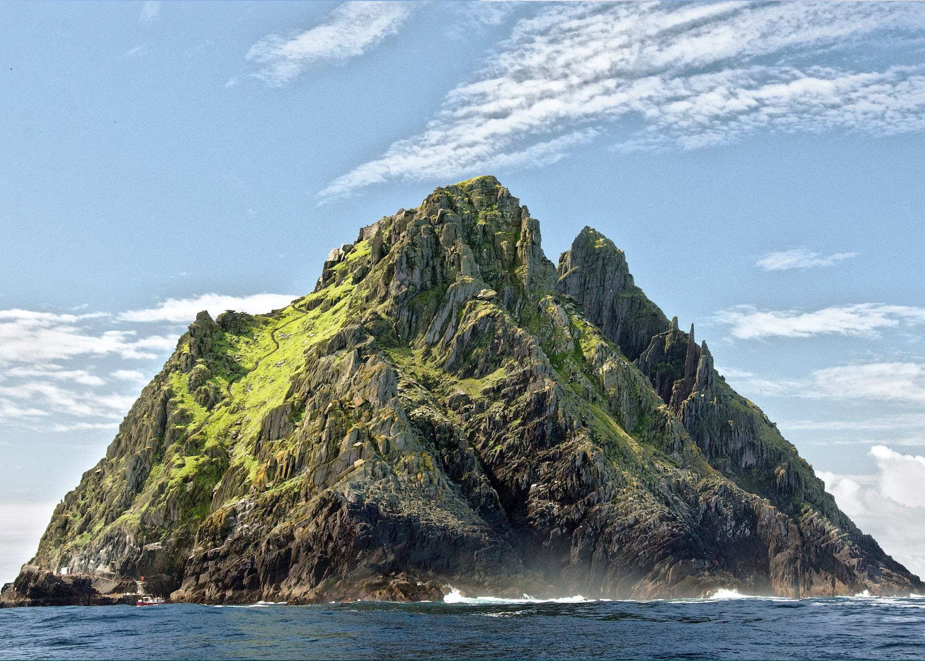 Dramatic rocky peak of Skellig Michael rising from the Atlantic Ocean, with moss-covered slopes, jagged stone formations, and sheer cliff faces against a blue sky with scattered clouds.
