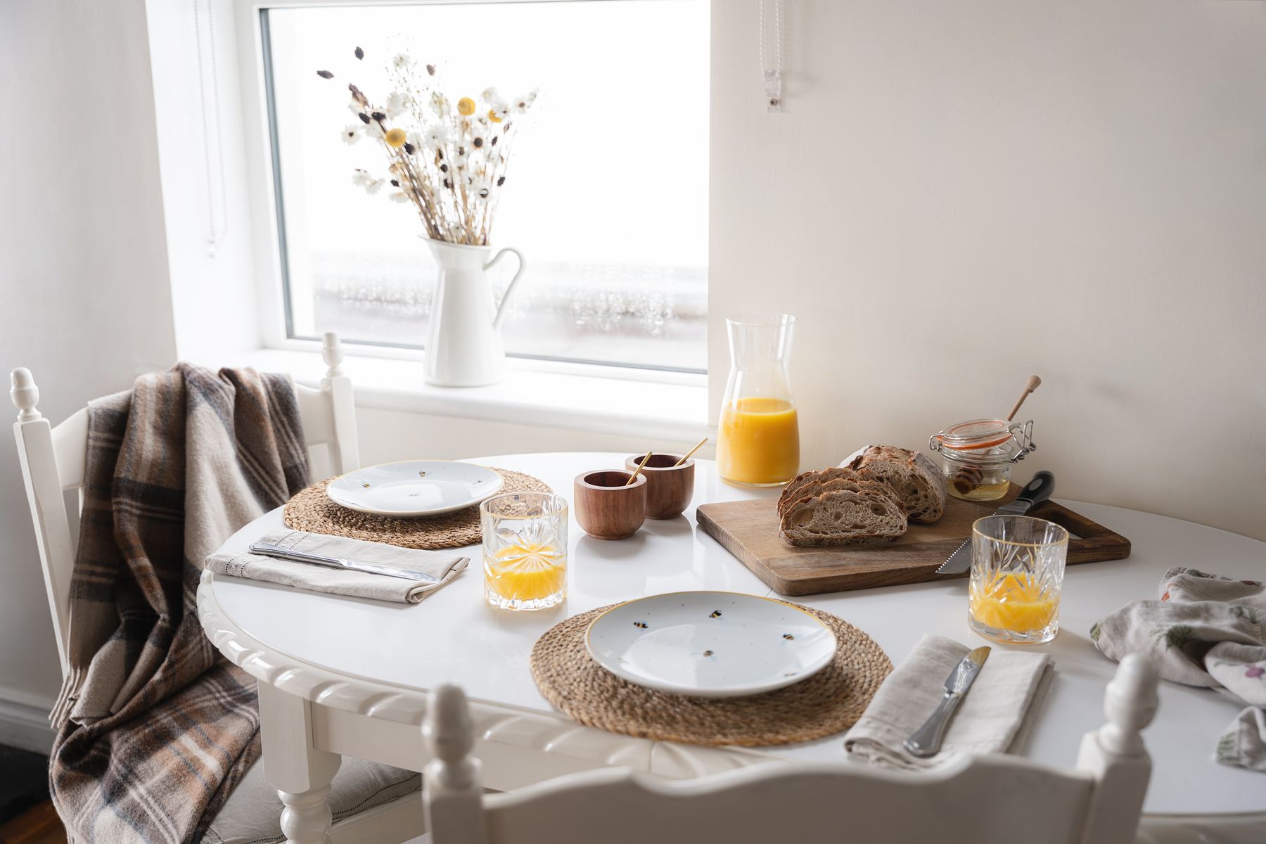 Close-up of a white breakfast table set for two with juice, bread, and honey, positioned by a bright window with a vase of dried flowers.