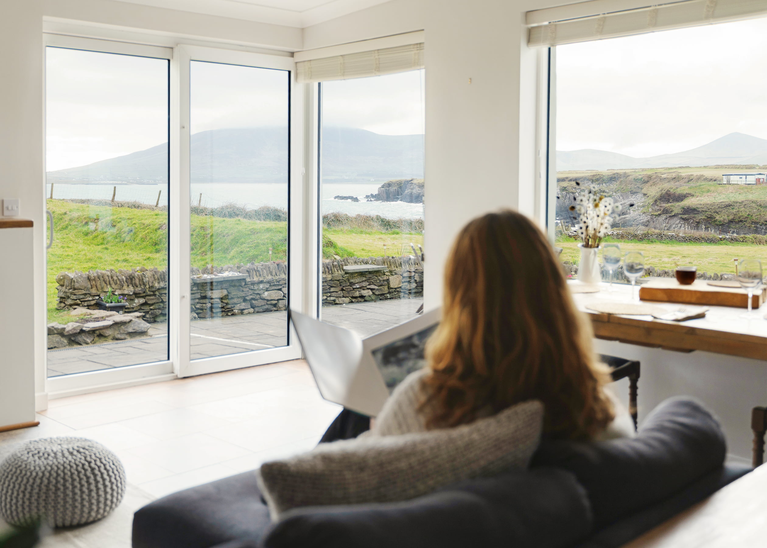 A guest sits on a sofa reading a book, facing large windows that look out onto grassy cliffs, stone walls, Ventry Bay, and Mount Eagle in the distance.