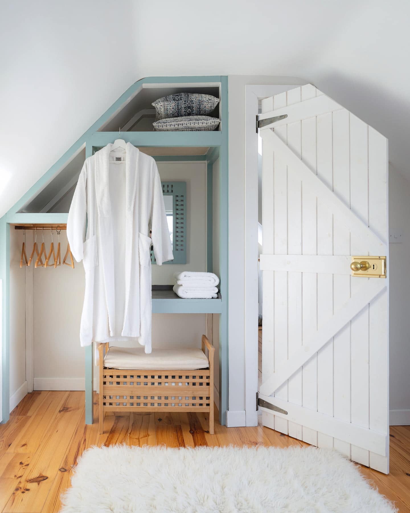 Charming bedroom nook at Ocean Blue, Sanctuary Cottages in Dingle, featuring a built-in wardrobe with soft robe, folded towels, natural wood bench, and fluffy rug on pine flooring beneath a sloped ceiling.