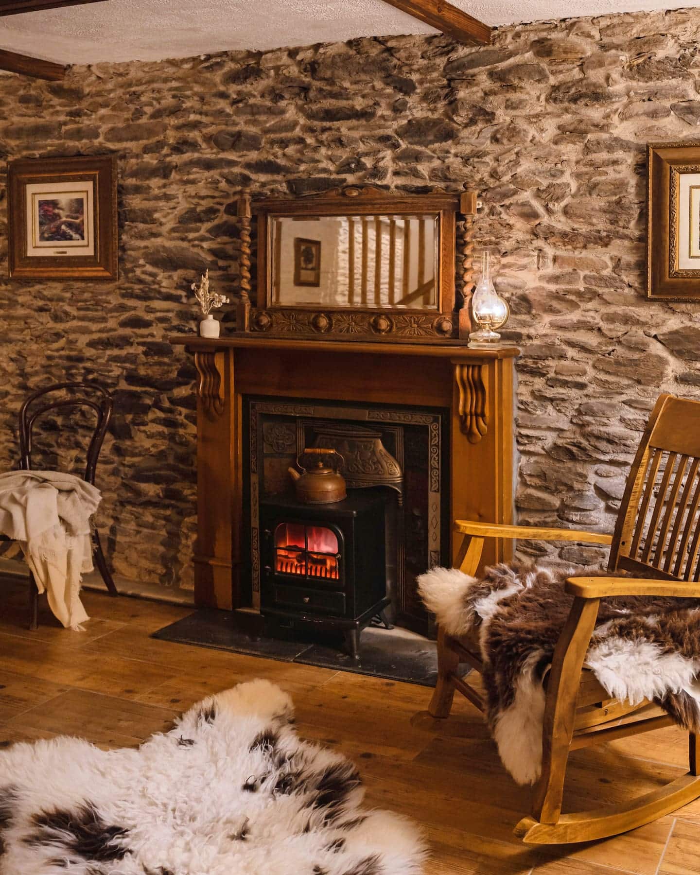 Warm and rustic living room at Pipers Cottage, featuring a traditional stone wall, wood-burning stove, antique mirror, and rocking chair with sheepskin throws.