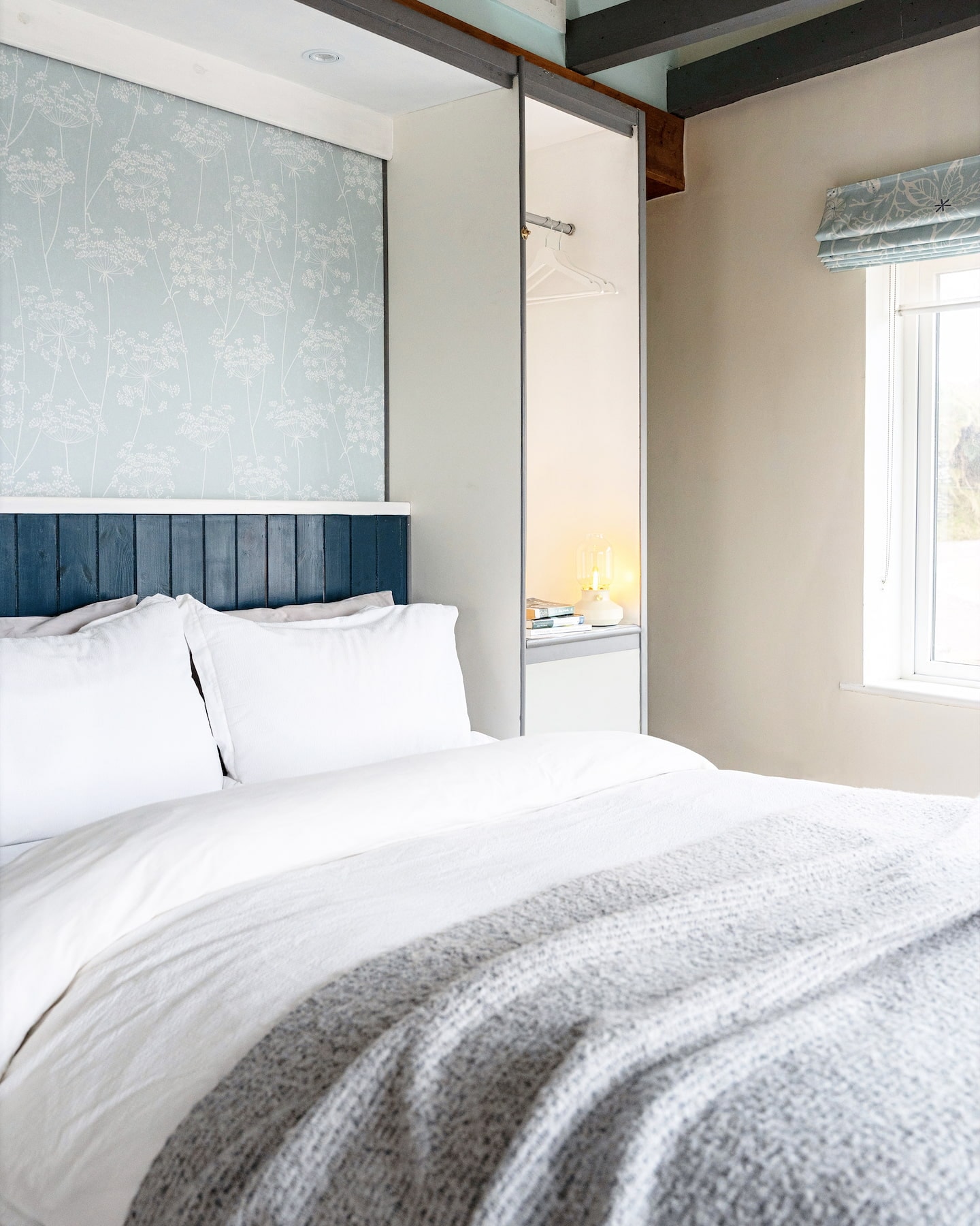Bright bedroom in The Studio at Sanctuary Cottages, Dingle, featuring a double bed with white linens, blue wood-panelled headboard, floral wallpaper, and built-in wardrobe with open shelving and hanging space.