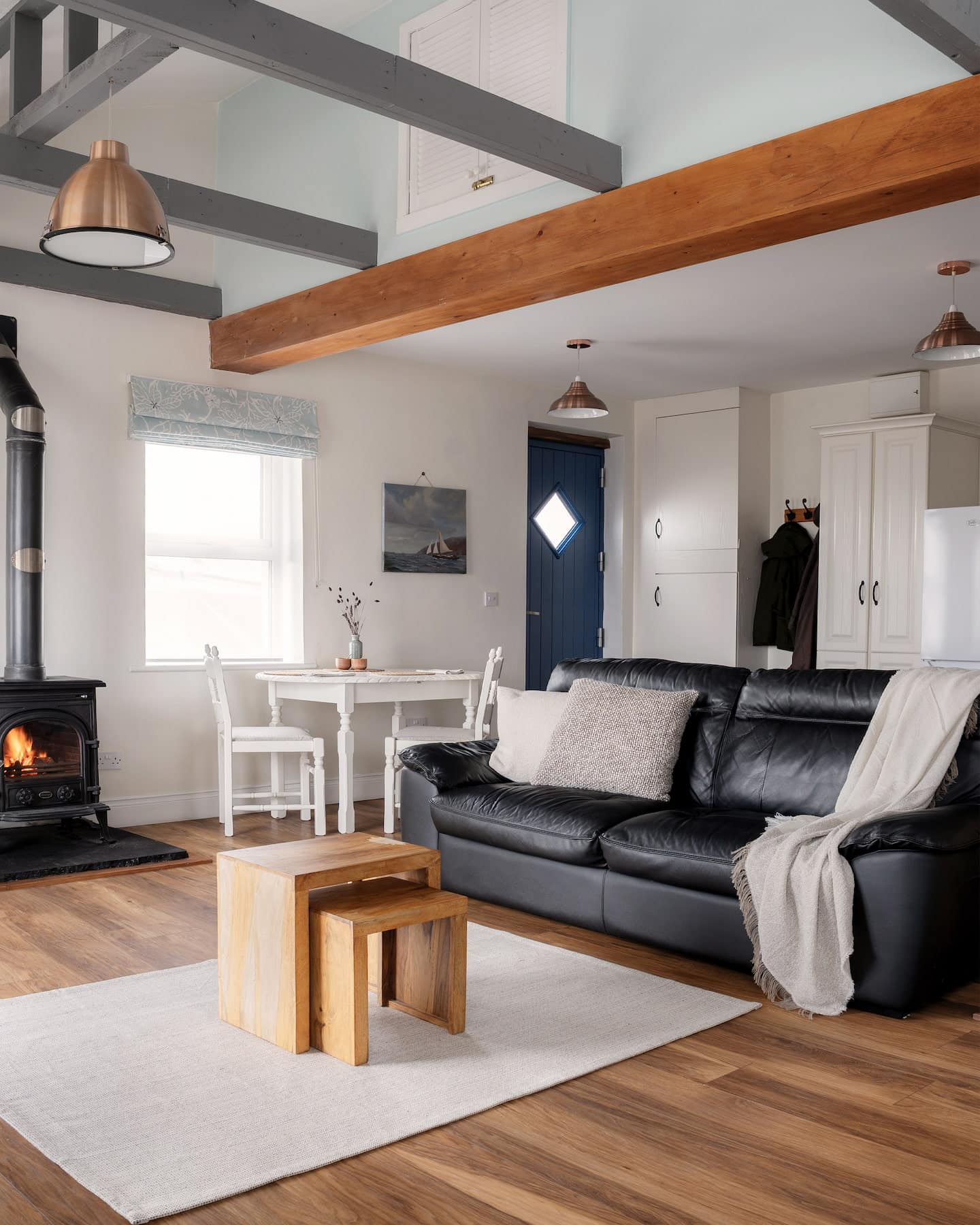 Spacious open-plan living area with a black leather sofa, wood-burning stove, dining nook, and natural light from multiple windows, accented by timber floors and vaulted ceiling beams.