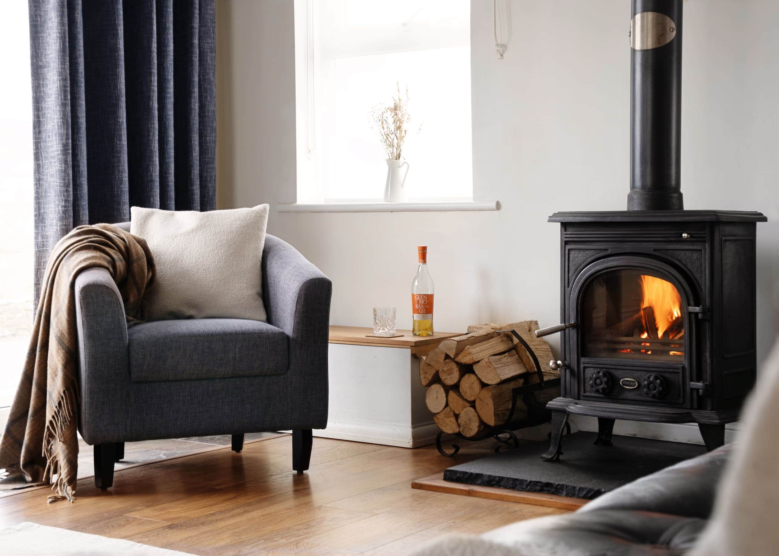 Cosy corner of The Studio at Sanctuary Cottages with a dark grey armchair, soft cushion, blanket, and a wood-burning stove set against a white wall with natural light from the window.